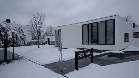 snow covering the garden of a modern white detached house for sale during winter, featuring large windows, a for sale sign and a cloudy sky