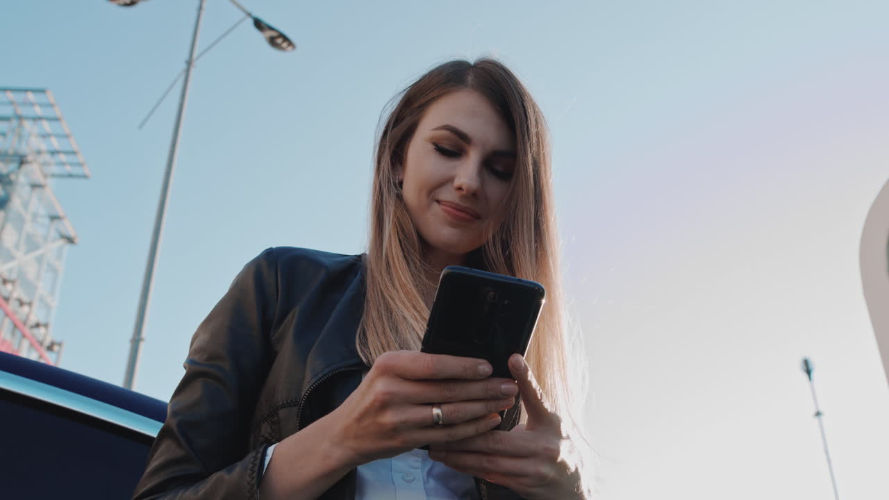 Giovane donna entusiasta che chatta al telefono in un'app di appuntamenti, si sente felice in piedi sulla strada vicino all'auto