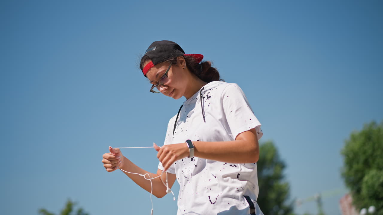 파란 하늘 아래에서 음악을 즐기는 십 대, 스플래터 티셔츠와 모자를 쓰고 이어폰을 테스트하며 미소짓는 모습, 나무와 구름이 배경에 있는 야외 전신 사진, 햇볕이 내리쬐는 편안한 분위기와 리듬감 있는 움직임