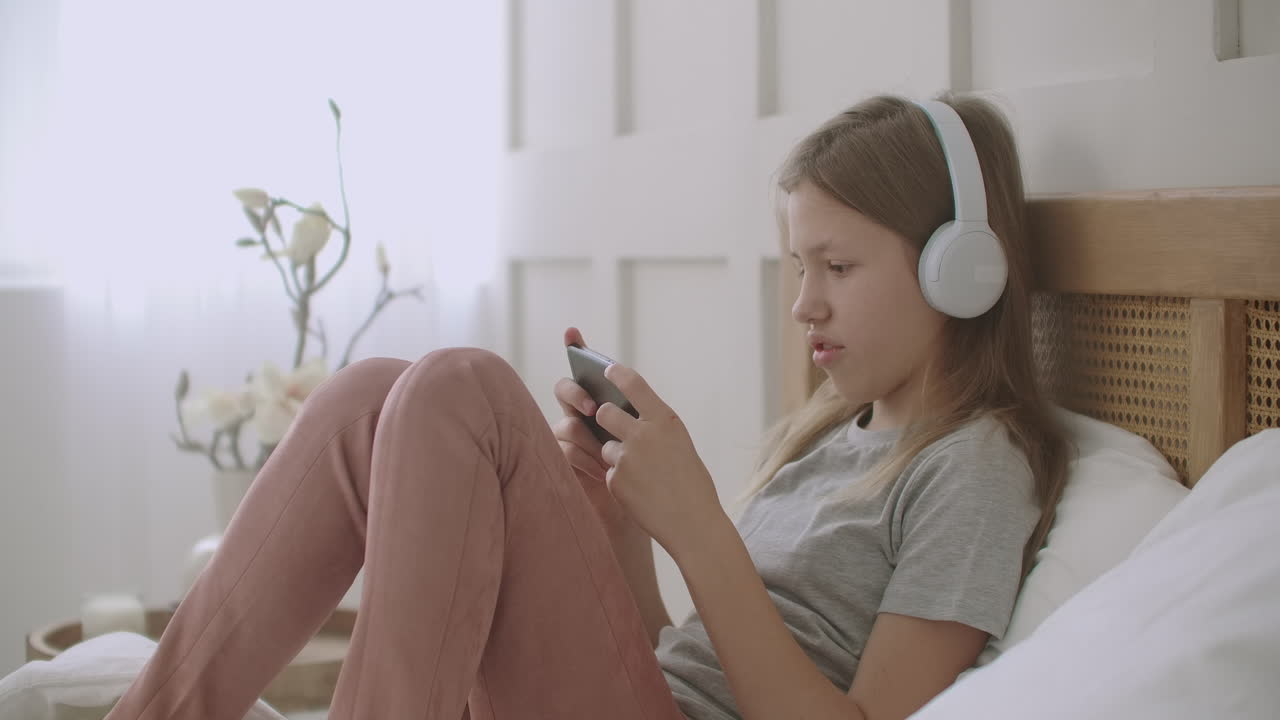 la niña de la escuela se queda en casa y juega juegos en el teléfono acostada en la cama escuchando música con auriculares