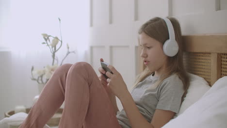 school girl is staying home and playing game in phone lying on bed listening to music by headphones