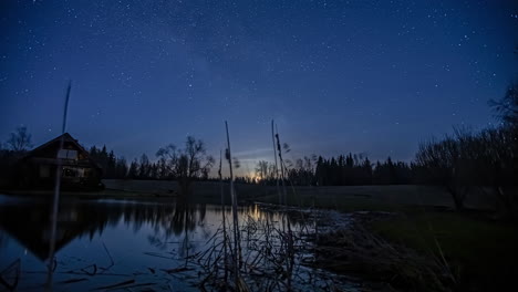 Vía-Láctea-Y-Luna-Moviéndose-Por-El-Cielo-Sobre-La-Cabaña-Junto-A-Un-Lago-Por-La-Noche