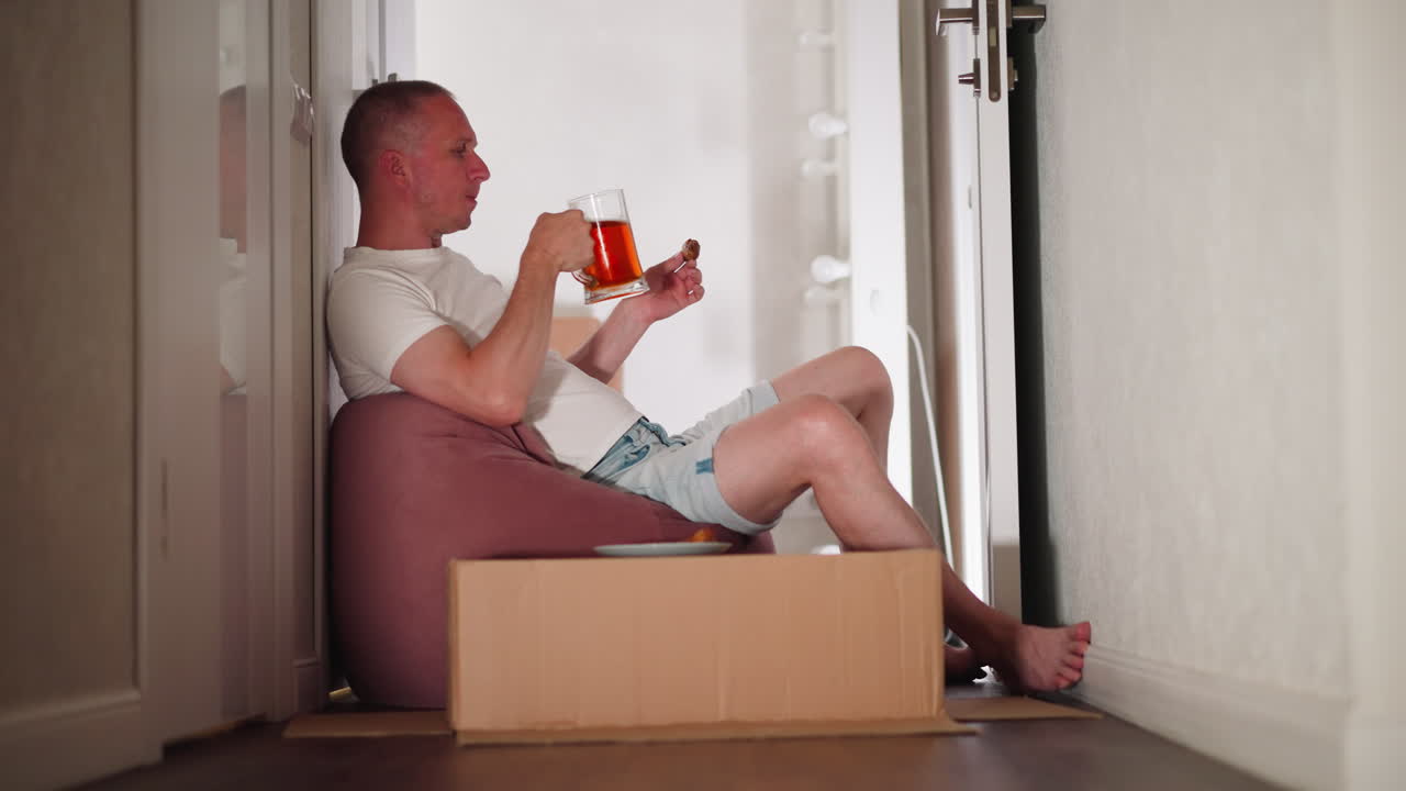 Young adult sitting relaxed in hallway drinking beer while holding meat in hand, places beer cup on carton used as makeshift table, casual home setting, peaceful atmosphere