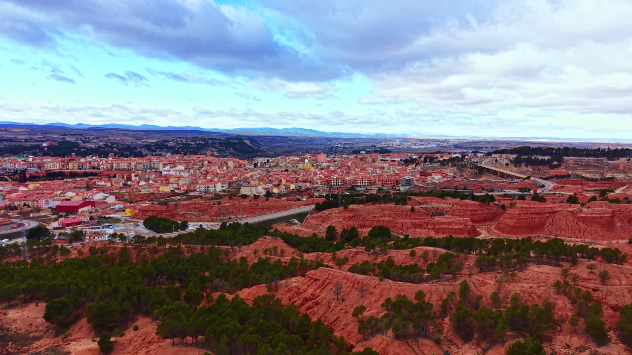 Veduta aerea di una città circondata da colline rosse e alberi verdi, ripresa nel pomeriggio con nuvole nel cielo e colline ondulate sullo sfondo.