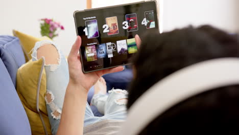 biracial man in headphones using tablet online lying on couch in living room, slow motion