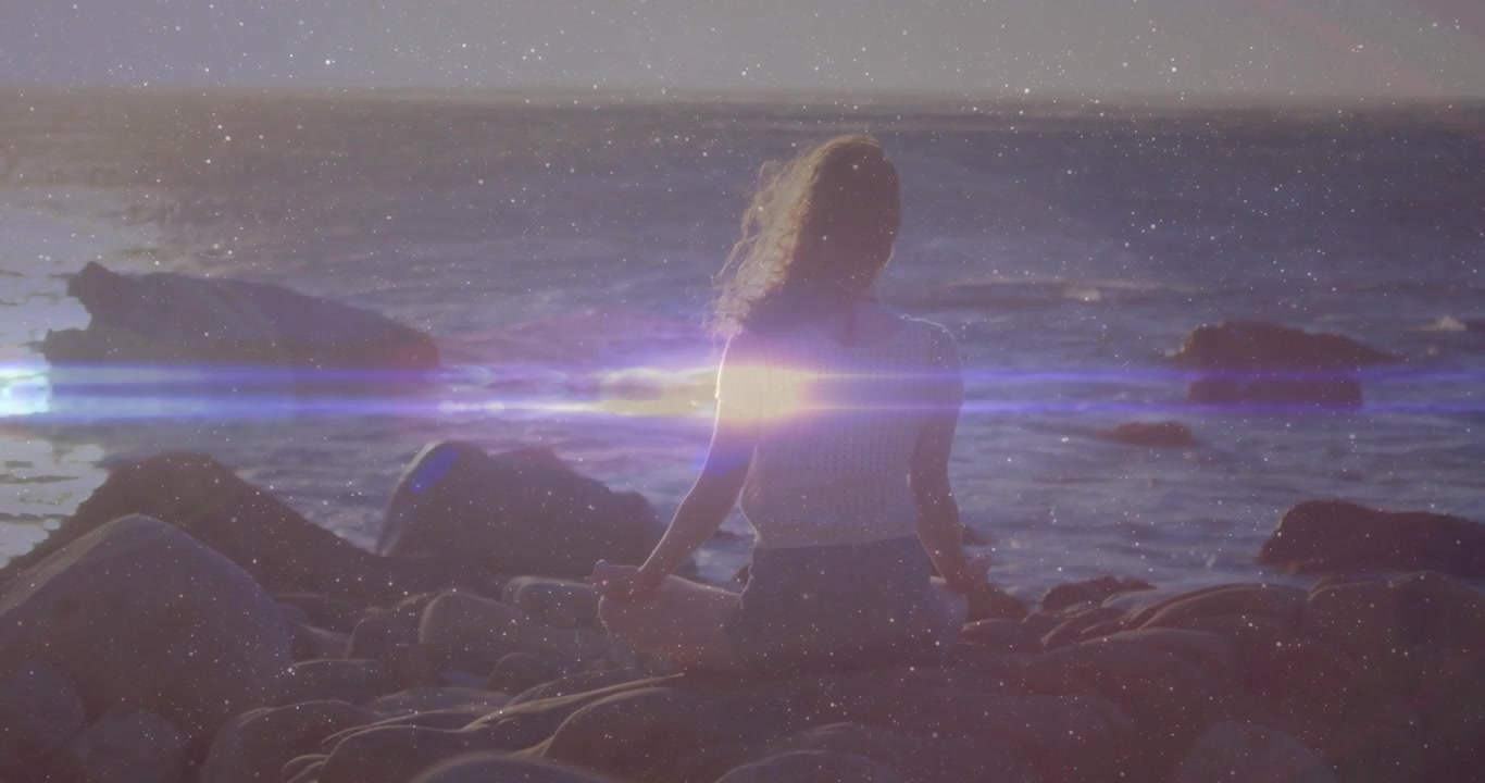 Kvinde mediterer på stenet kystlinje for sundhed, solen nærmer sig horisonten, hvilket forårsager en rejsende linseflare.
