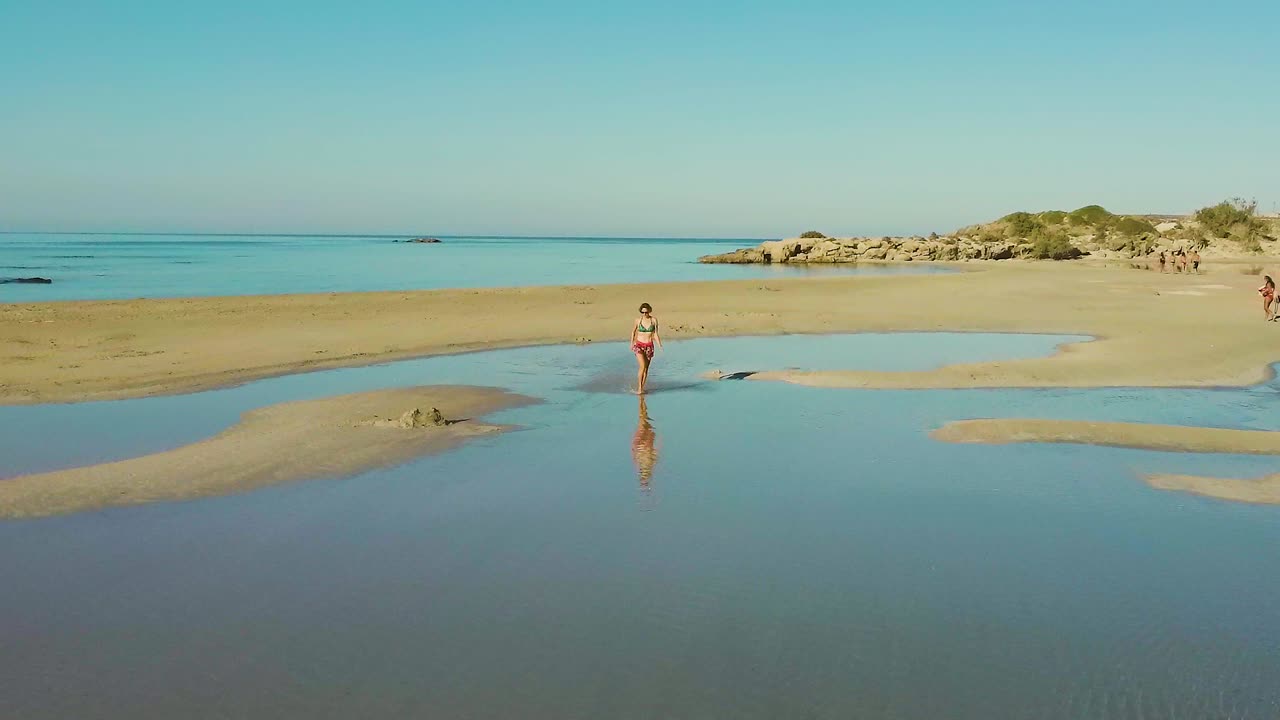 low flying drone aerial view of woman running in empty elafonissi beach lagoon reflection of blue clear sky