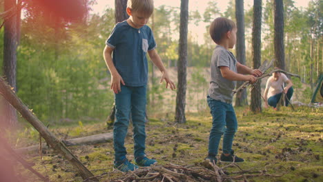 Two-boys-collect-sticks-in-the-woods-for-a-large-family-campfire-against-the-backdrop-of-a-tent-and-a-lake.-Family-is-going-to-light-a-fire-for-camping-and-frying-marshmallows