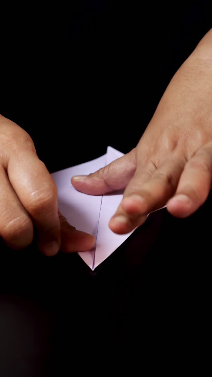 Adult hands carefully fold white paper on dark surface, demonstrating origami preparation with soft lighting