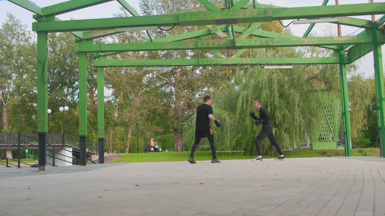 To hvite menn trener under en pergola i en park, treneren demonstrerer forsvarsteknikker mens studenten øver på blokkeringer og motangrep, skarpt fotarbeid på asfaltert grunn med grønn metallramme og piletrær