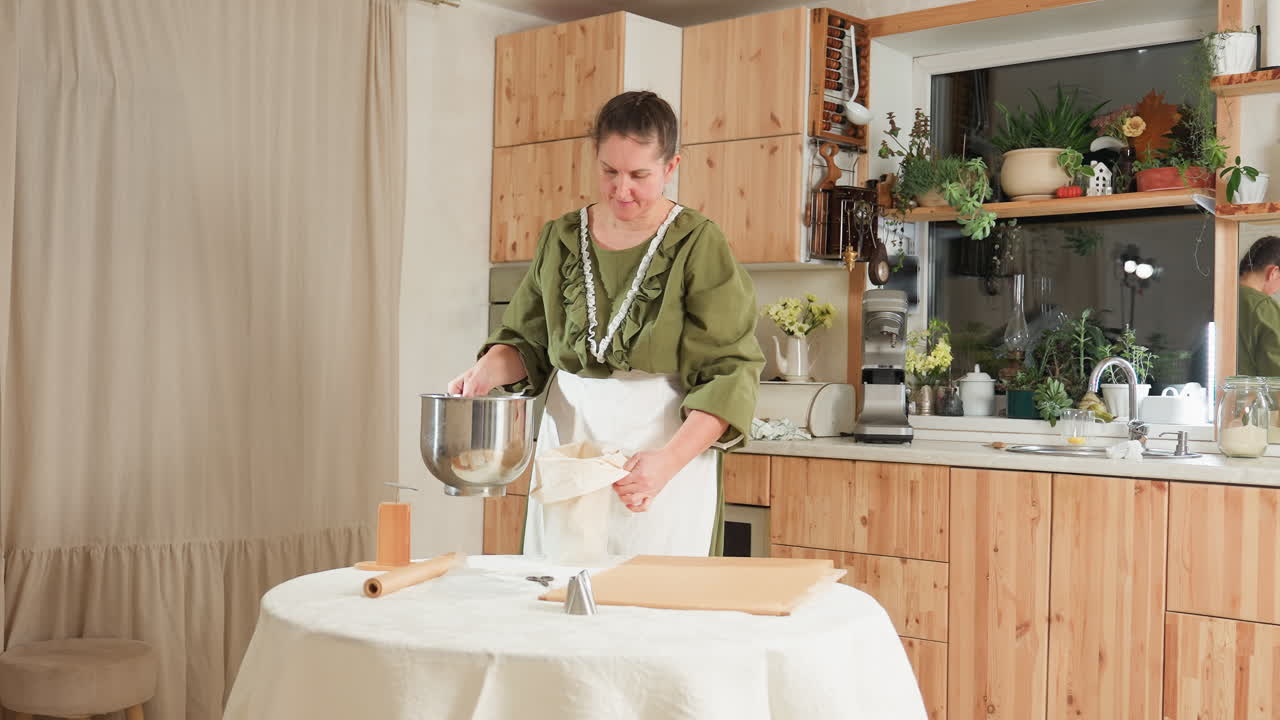 Baker arranges cloth neatly inside jug while preparing for baking, then lifts metal bowl containing flour and picks up mixer tool from table in well-organized home kitchen filled with natural light