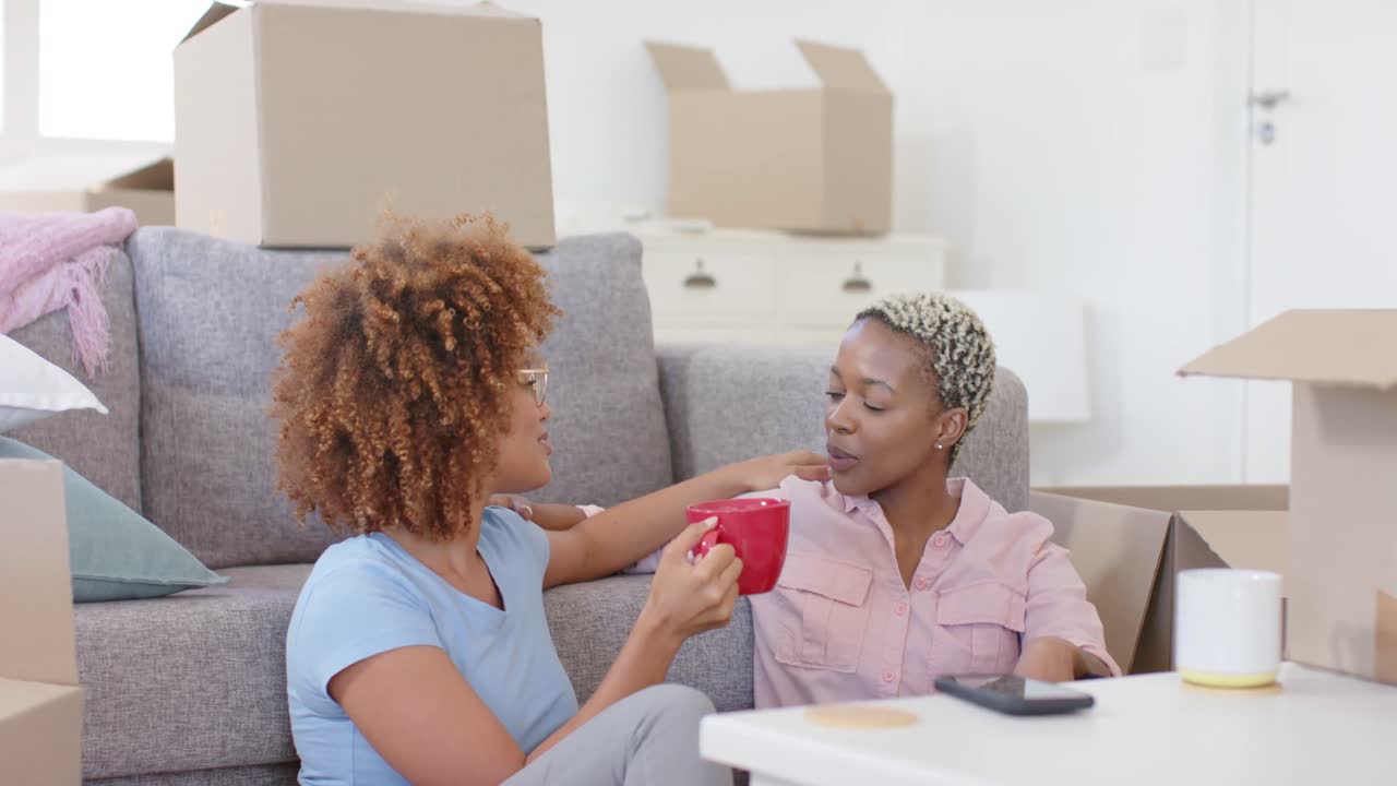 Happy diverse female lesbian couple with boxes moving house and drinking coffee in slow motion