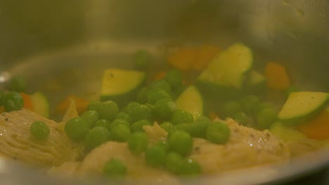 The-cook-tops-fresh-green-peas-over-chicken-and-zucchini-in-the-steel-pot,-making-pasta-salad,-handheld-close-up-shot