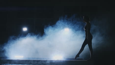 Side-view-of-a-female-gymnast-doing-split-handstand-on-balance-beam-against-black-background.