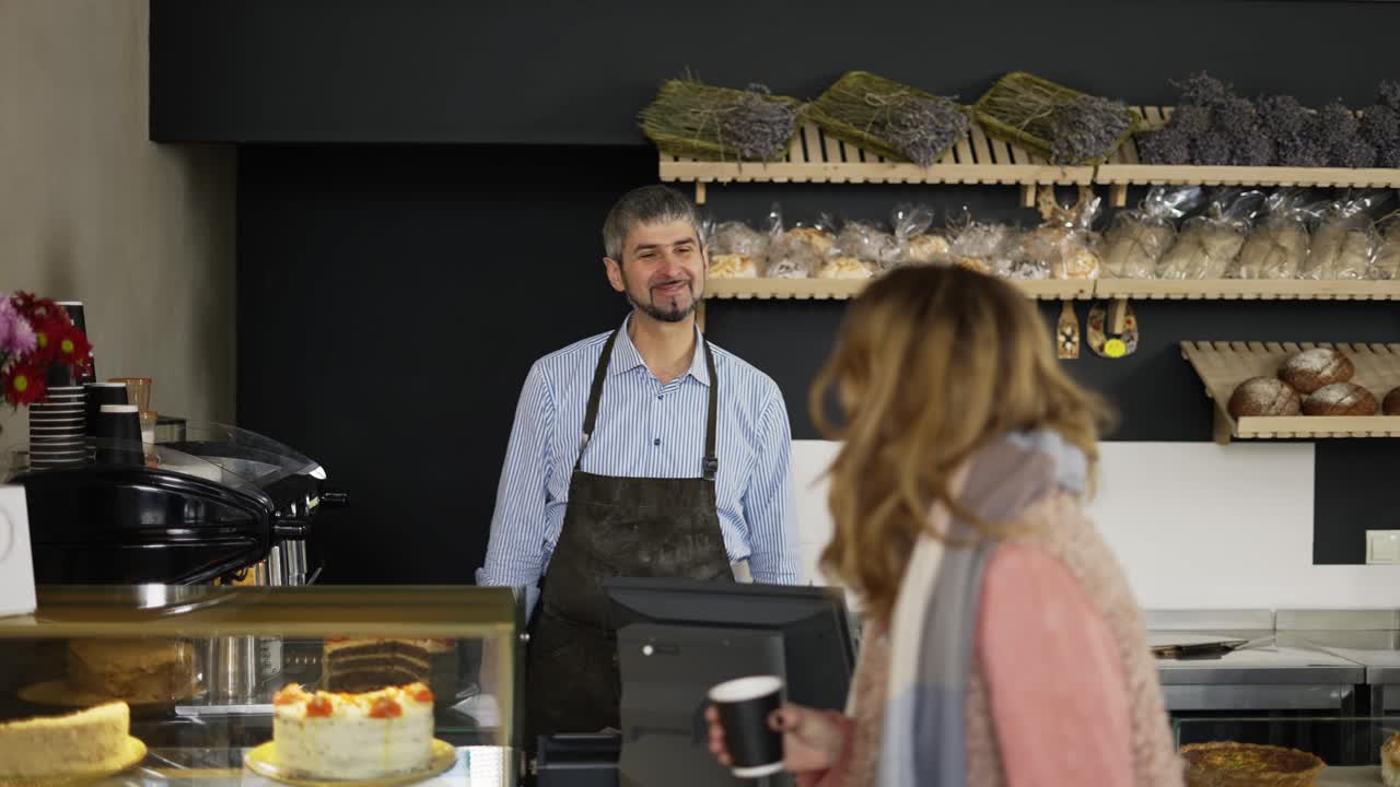 atractivo barista masculino en delantal negro dando taza de papel café para llevar al cliente femenino. interior, panadería