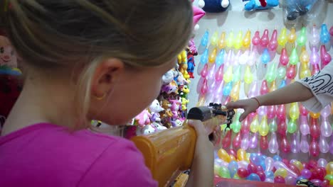 Young-Caucasian-girl-shooting-rubber-bullet-gun-at-rows-of-colorful-balloons-to-win-prizes-at-shooting-range