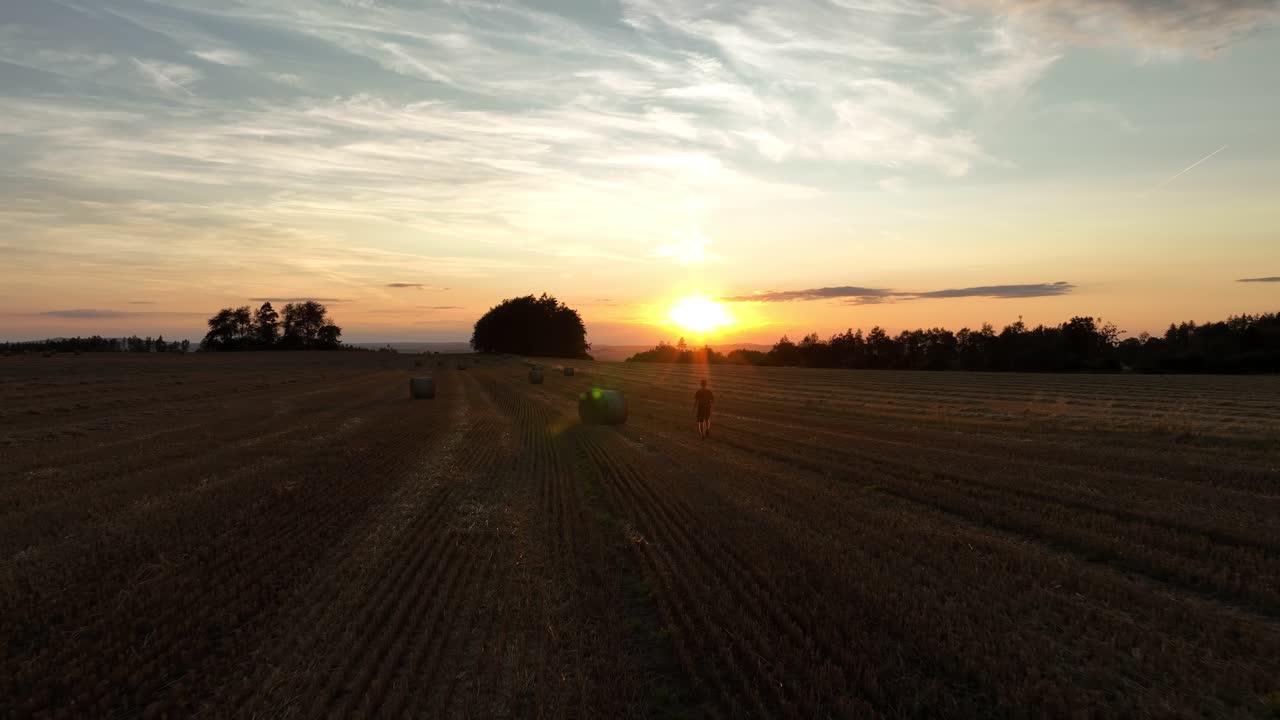 un hombre solitario caminando a través de un campo cortado con balas de heno, mirando a la distancia al atardecer, disparo de drones, cámara lenta