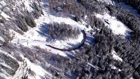 Aerial-view-of-the-Bernina-Express-panorama-train-going-through-a-snow-covered-mountain-winter-landscape-with-forests-on-a-sunny-day-in-Alp-Grum,-Switzerland