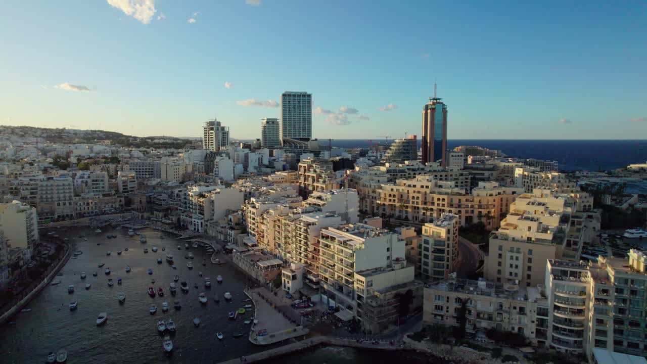 Drone pans then advances over St. Julian’s Bay in Malta, showing boats, hotels, and buildings in morning light