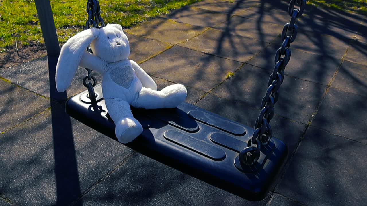 Coniglietto di peluche solitario che dondola lentamente su un'altalena di un parco giochi deserto, una scena toccante di ricordi d'infanzia dimenticati e solitudine in una giornata di sole con lunghe ombre degli alberi.