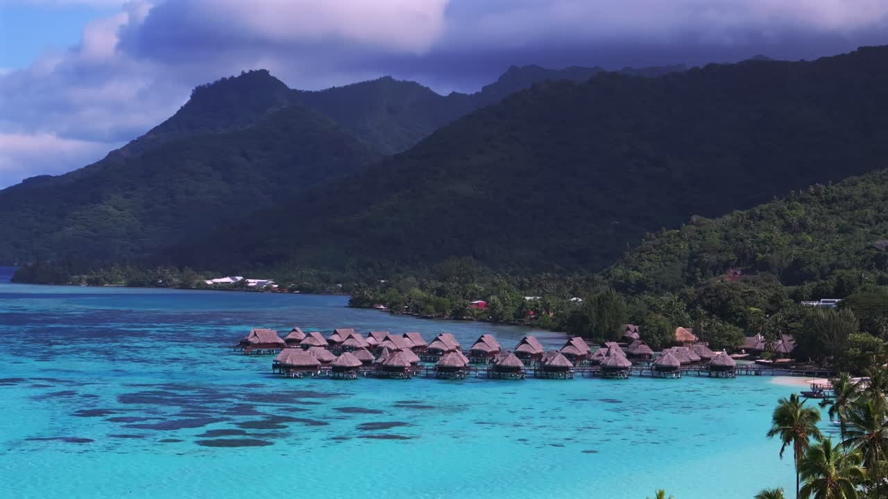 Droneudsigt over Mooreas revomkransede lagune i Fransk Polynesien, hvor lyst turkisblåt vand møder Ta'ahiamanu Strand over vandbungalows under frodige vulkanske bjerge, lave skyer cirkler til højre.