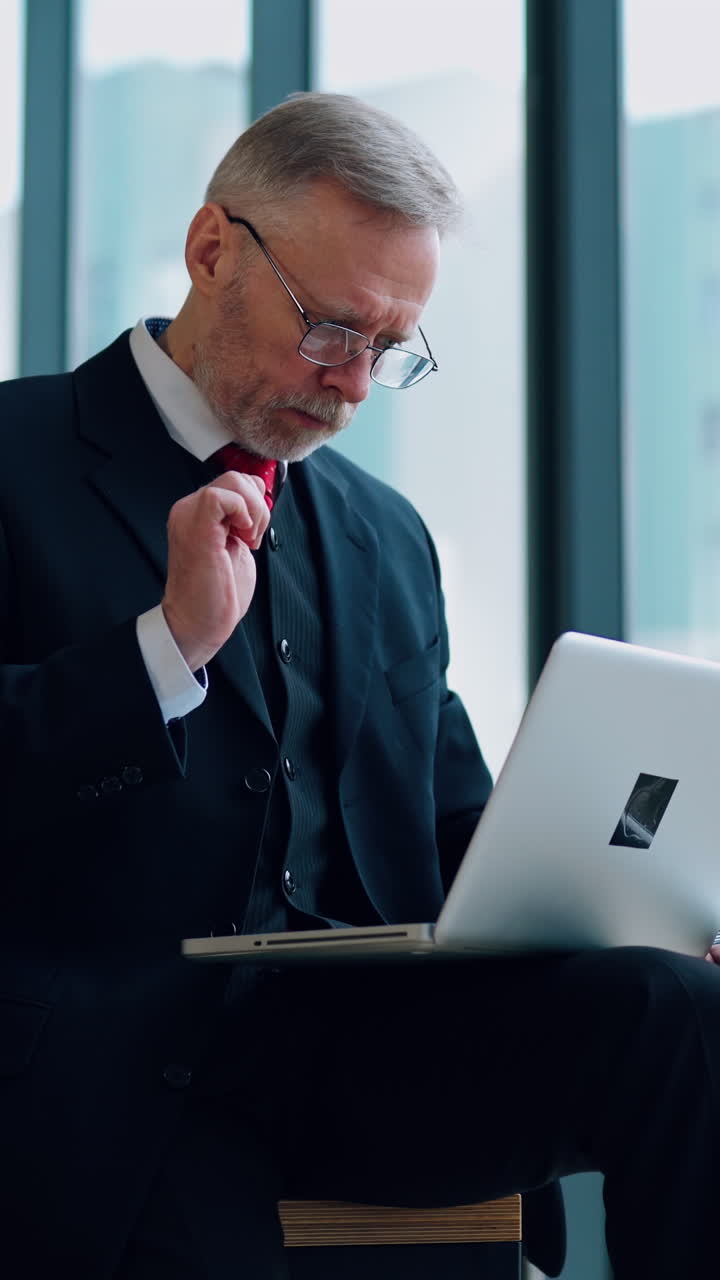 Senior businessman in suit working near office window. Elderly elegant man is typing on a laptop while sitting near the window. Business concept. Vertical video