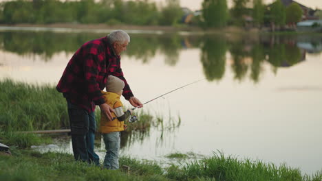 Descanse-En-La-Naturaleza,-El-Abuelo-Y-El-Nieto-Están-Pescando-Juntos-En-La-Hermosa-Orilla-Del-Lago-Por-La-Mañana