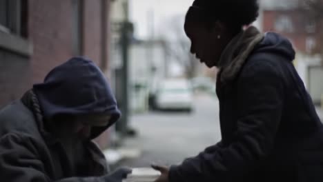 on a chilly evening, a woman offers food to a homeless man sitting on the street. the atmosphere is reflective as passersby move through the urban landscape.