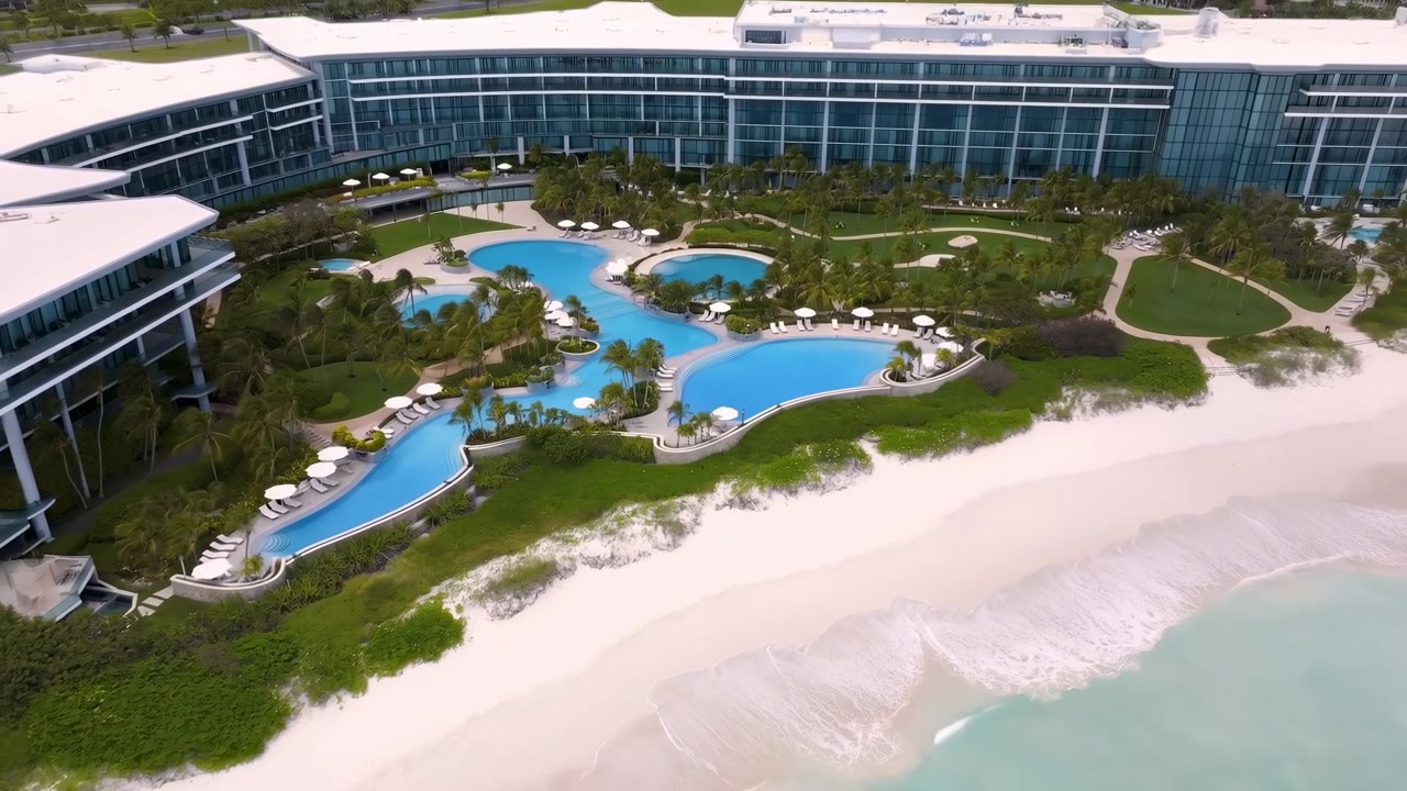 Vista diurna dall'alto di un resort a forma di mezzaluna sul lungomare, con diverse piscine a laguna interconnesse di colore blu acqua, ombrelloni e lettini bianchi, palma.