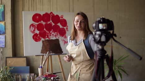 female caucasian mature blogger recording video in workshop - drawing pink flower on canvas, explaining details on camera. slow motion