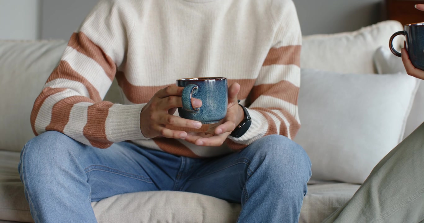Coppia afroamericana seduta a parlare sul divano, l'uomo tiene una tazza blu, cambia la presa e mostra l'orologio.