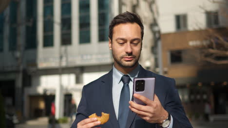 Cheerful-Bearded-Business-Man-Using-Phone-and-Eating-Wholegrain-Donut-Commuting-to-Office-in-Urban-Background