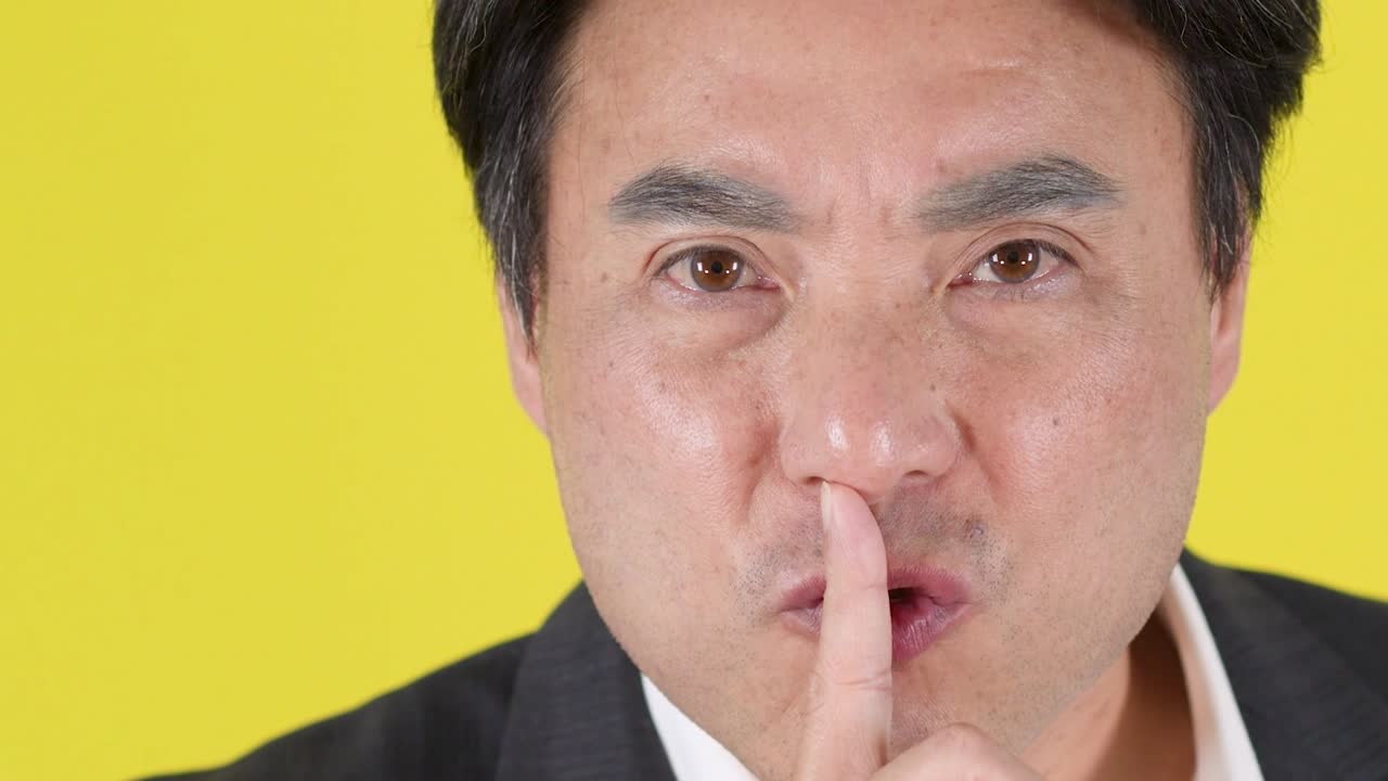 スーツを着た真剣なアジア人男性が、唇に指を当てて静かにするよう合図している。