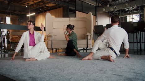 A-girl-and-two-guys-in-white-and-green-business-suits-are-sitting-on-a-rug-in-the-office-and-doing-a-Spine-Twisting-Pose.-Yoga-classes-during-a-break-from-office-work