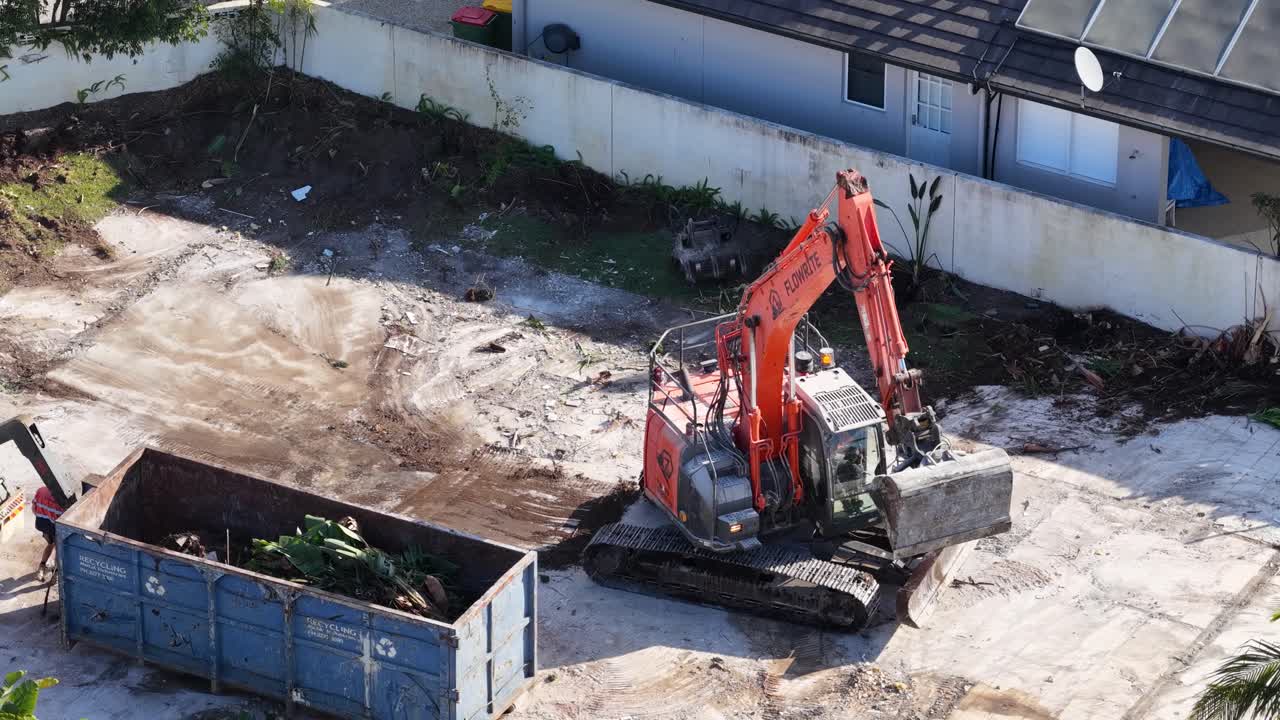 An orange excavator transfers demolition debris into a large blue dumpster at a cleared construction site in daylight, viewed from above with steady camera