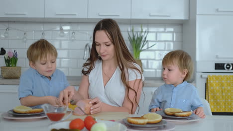 Dos-Niños-Pequeños-Ayudan-A-Mamá-En-La-Cocina-A-Preparar-Hamburguesas-Cortando-Verduras,-Queso-Y-Salchichas