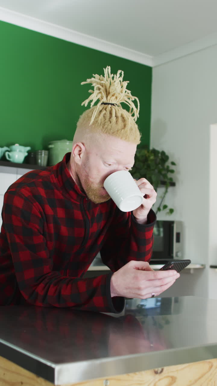 hombre albino se inclina en la isla de la cocina, toma un sorbo de café, usa el teléfono inteligente