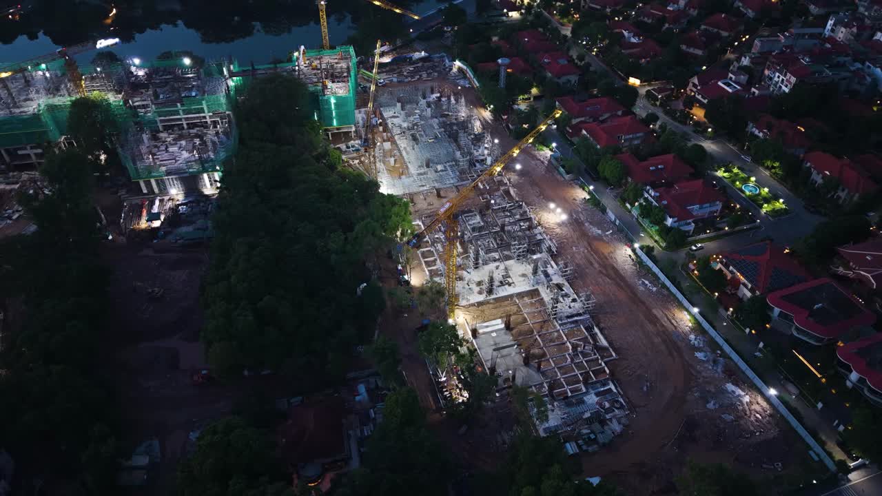 Aerial night view of an illuminated construction site in Tay Ho, Hanoi, Vietnam, showing cranes, building frameworks, and a nearby residential area beside West Lake