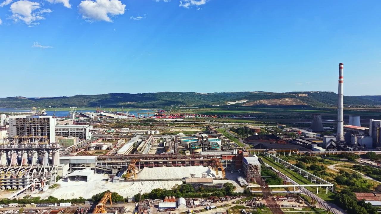 Vista di un'area industriale con fabbriche e ciminiere vicino a un fiume e colline in una giornata di sole.