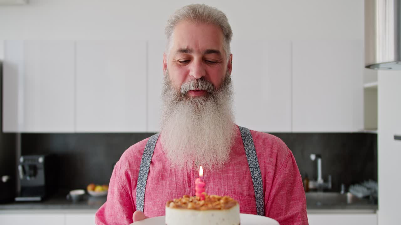 retrato de un hombre mayor feliz con cabello gris con una barba exuberante en una camisa rosa que sostiene un pastel con una vela encendida delante de él y lo sopla en honor a su cumpleaños en una cocina moderna