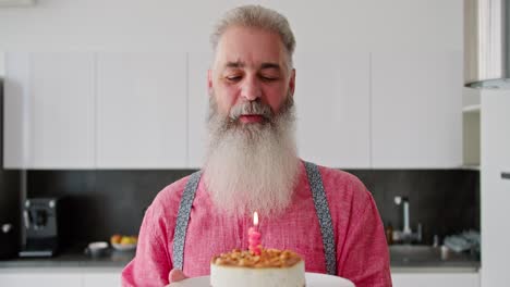 Portrait-of-a-happy-elderly-man-with-gray-hair-with-a-lush-beard-in-a-pink-shirt-who-holds-a-cake-with-a-burning-candle-in-front-of-him-and-blows-it-out-in-honor-of-his-birthday-in-a-modern-kitchen