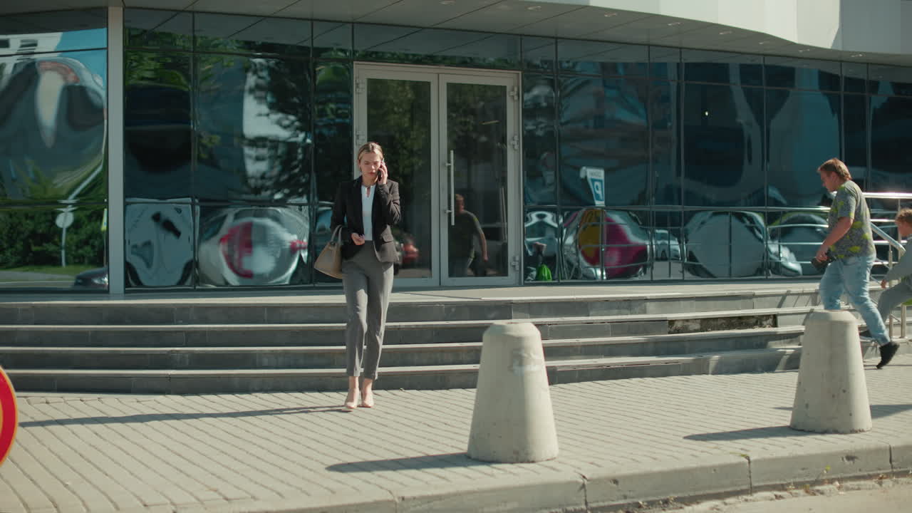 Young businesswoman in formal outfit on phone call descending stairs outside modern office building with glass reflections of pedestrians and car passing by