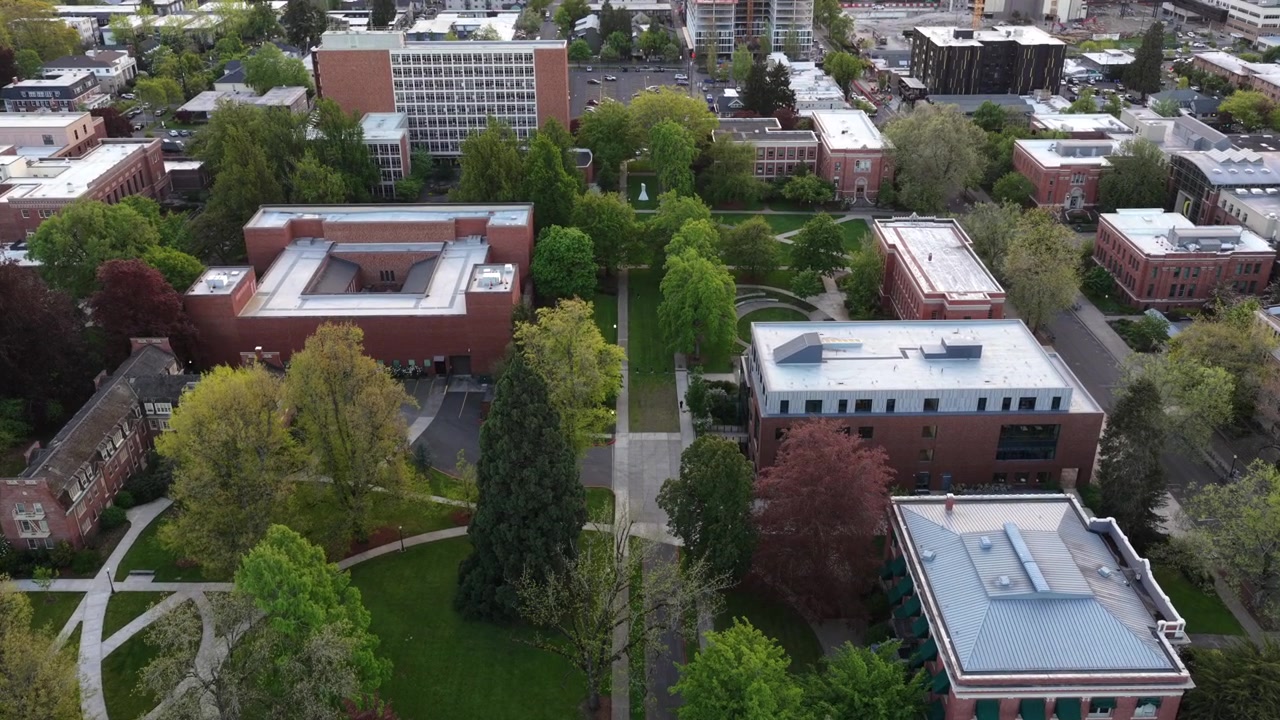 Oregon Üniversitesi kampüs meydanı; kırmızı tuğlalı binalar, kavisli yollar, gür bahar ağaçları, yeşil çimler ve bulutlu gökyüzünün altında çevresindeki Eugene şehir manzarası. Oregon, ABD. Havadan genel çekim.