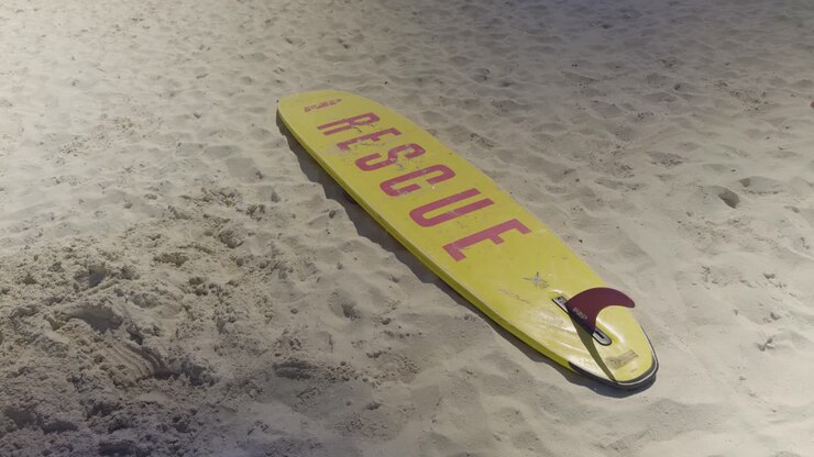 A yellow lifeguard rescue board lies on soft, white sand under bright daylight. The bold red “RESCUE” text is clearly visible, evoking safety, readiness, and coastal emergency services.