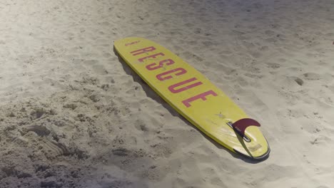 a yellow lifeguard rescue board lies on soft, white sand under bright daylight. the bold red “rescue” text is clearly visible, evoking safety, readiness, and coastal emergency services.