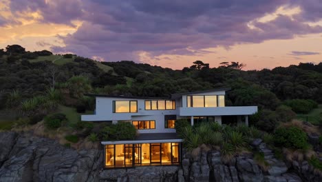 luminous contemporary residence perched on cliff edge, radiating golden warmth against forest backdrop, embodying serene coastal architectural elegance amid verdant landscape