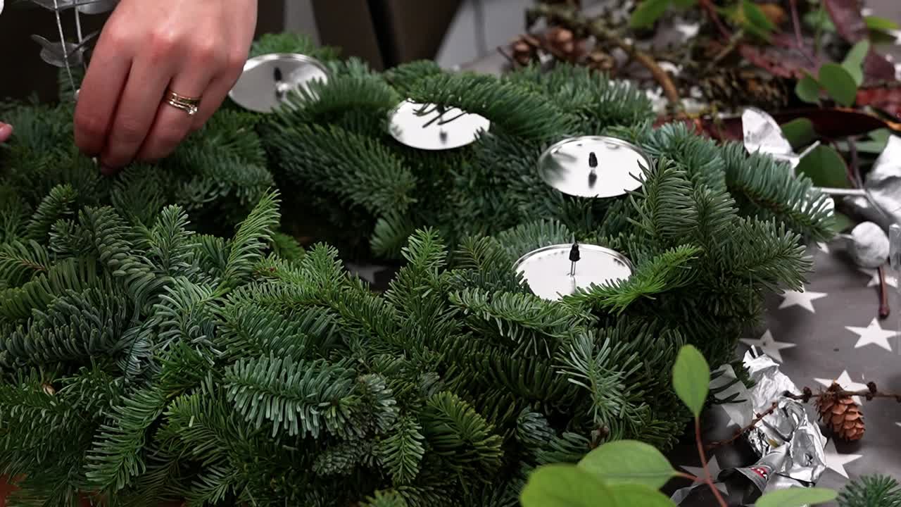 Raffinato primo piano di mani che decorano una lussureggiante ghirlanda natalizia con candele, che mette in mostra tecniche dettagliate di composizione floreale in un ambiente accogliente. Perfetto per trarre ispirazione per design festivi.