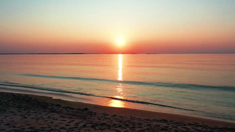 Vista-Del-Hermoso-Amanecer-En-El-Océano-Mientras-Camina-En-La-Playa-Con-Un-Sol-Brillante-Que-Proyecta-Coloridos-Reflejos-Rojos,-Naranjas,-Morados-Y-Amarillos-Sobre-Las-Tranquilas-Olas-Del-Mar-Con-Un-Hermoso-Cielo