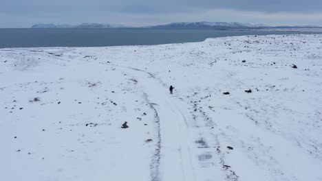 Viajero-De-Aventuras-Solitario-Caminando-Por-Un-Camino-Nevado-Hacia-Los-Acantilados-Costeros-En-Un-Día-Nublado,-Aéreo