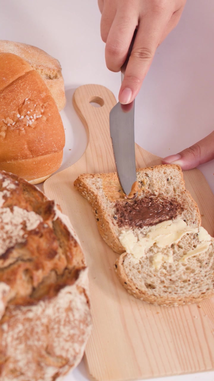 Mano unta mantequilla de cacahuete cremosa sobre tostada multigrano rodeada de pan de masa madre y baguettes crujientes